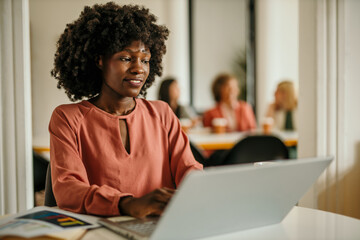 Shot of an adorable african-american businesswoman using a laptop and making notes on a clipboard inside of the office.