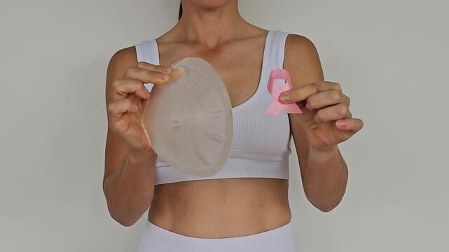 A woman holds a silicone breast implant and a pink ribbon to symbolize breast cancer awareness. Breast cancer and breast plastic surgery. 