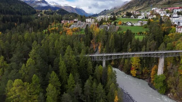 Aerial flyover bridge over River Inn with walking tourist in Scoul Village, Switzerland,. Snowy mountains range and luxury houses and apartment with river view. Establishing shot.