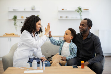 African female doctor gives high five to young boy while sitting with father at home. Warm interaction promotes trust and comfort in home healthcare setting.