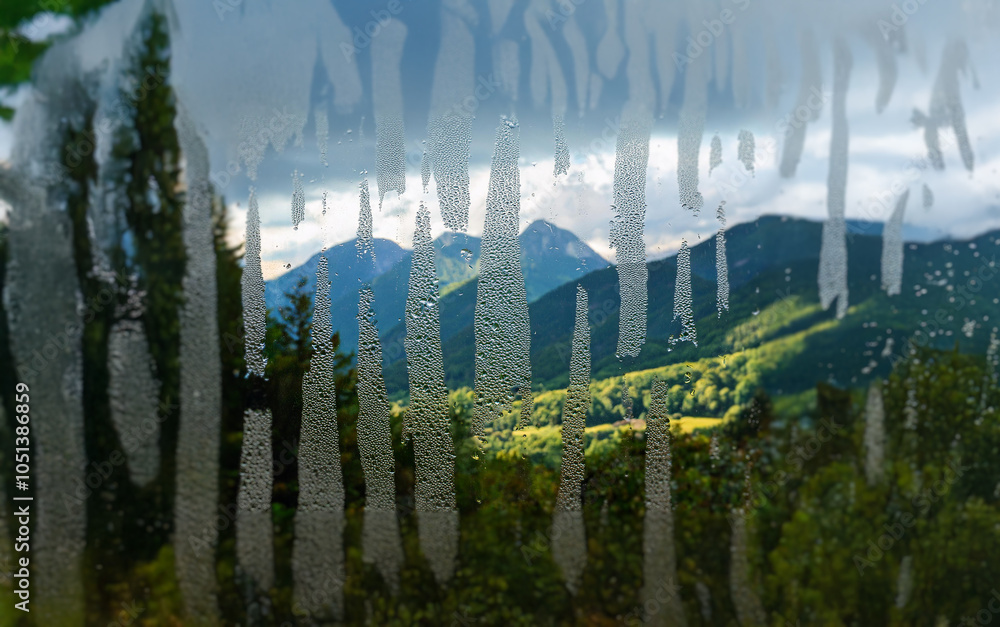 Poster A scenic mountain range with lush green slopes and a cloudy sky is seen through a window with streaks of raindrops.