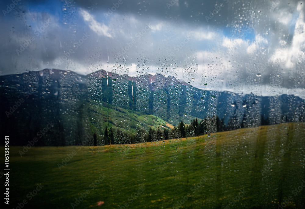 Poster A view of a grassy field with mountains in the background, seen through a window with rain droplets.