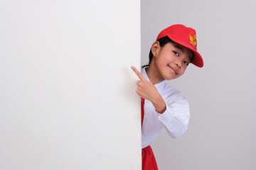 Indonesian school student peeking behind wall and pointing on it with happy face