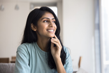 Happy daydreaming young Indian woman posing at home, looking away with toothy smile, touching chin, facial skin, sitting on couch, thinking, dreaming, enjoying homey comfort