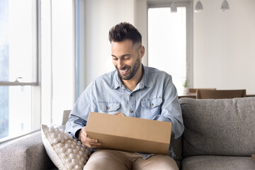 Cheerful Latin customer man unpacking received parcel, opening small cardboard box on home sofa,...