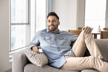 Happy attractive Hispanic guy watching TV, resting on soft comfortable home couch, enjoying football match, movie, sitcom show, comedy, holding remote control, smiling, looking away