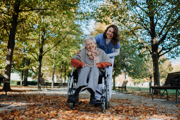 Disabled elderly woman in wheelchair on walk in the park during warm autumn day. Young nurse...