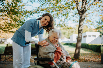 Disabled elderly woman in wheelchair on walk in the park during warm autumn day. Young nurse...