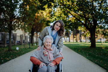 Fototapeta premium Disabled elderly woman in wheelchair on walk in the park during warm autumn day. Young nurse pushing wheelchair, talking with senior woman.
