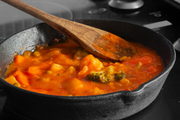 Homemade vegetable soup in a cast iron frying pan with a wooden spoon cooking on an electric hob...
