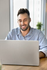 Fototapeta premium Positive handsome freelance professional man in casual working on online project at laptop, typing, smiling, browsing Internet, social media, sitting at workplace table in home office. Vertical shot