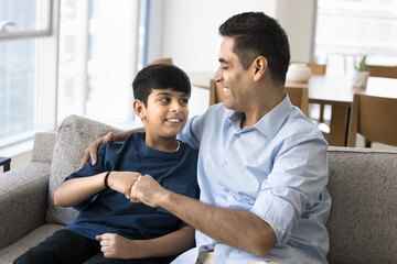 Happy Indian dad and teenage son making fist bump friends gesture, talking and laughing on home...