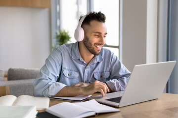 Happy adult Latin student man in headphones studying online at laptop and open books on table, watching lecture, webinar on computer, speaking on conference chat, using technology for education