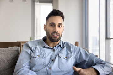 Obraz premium Handsome Latin blogger man filming self video for vlog at home. Influencer looking at camera, speaking, sitting on home sofa, giving webinar, lecture, online class, talking on video conference call