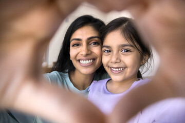 Happy beautiful Indian mom and sweet daughter kid looking at camera through finger heart shaped frame, smiling, showing hand gesture symbolizing love, family, childcare, friendship