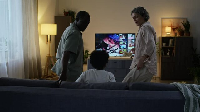 Rear view of diverse family coming to living room to watch movies on streaming service together in evening, active boy jumping onto sofa and parents with popcorn taking seat in front of TV