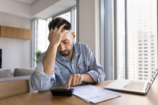 Worried tired freelance businessman getting financial problems, using calculator at home office workplace, touching head in frustration, finding bankruptcy risk, making difficult decision