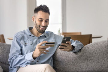 Happy satisfied Latin bank customer man shopping online, using mobile phone and credit card for...