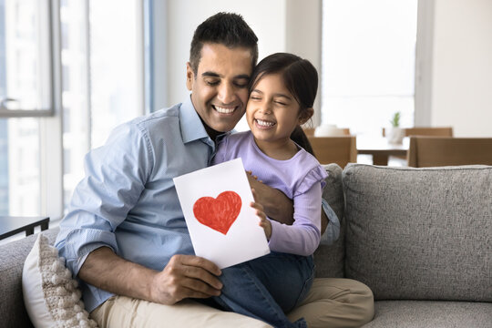 Happy excited Indian dad holding handmade greeting card and daughter kid in arms, celebrating fathers day, birthday, hugging little girl with closed eyes, smiling, laughing