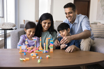 Happy Indian mom and dad helping kids to build toy towers, constructing colorful pieces, enjoying...