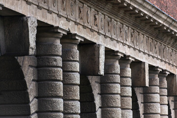 A grand colonnade with robust, textured stone pillars stands in quiet strength. The repetitive arches create a rhythmic architectural pattern against a high brick wall, surrounded by greenery.