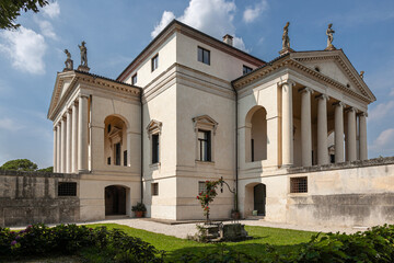This grand villa showcases Palladian architecture with a stately colonnade, sculpted statues on the roofline, and arched entrances. A serene garden and central well enhance the classical ambiance.
