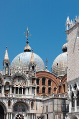 A masterpiece of Venetian architecture shines under the blue sky. St. Mark's Basilica showcases its elaborate façade with intricate arches, statues, and iconic Byzantine domes, blending mosaic and mar