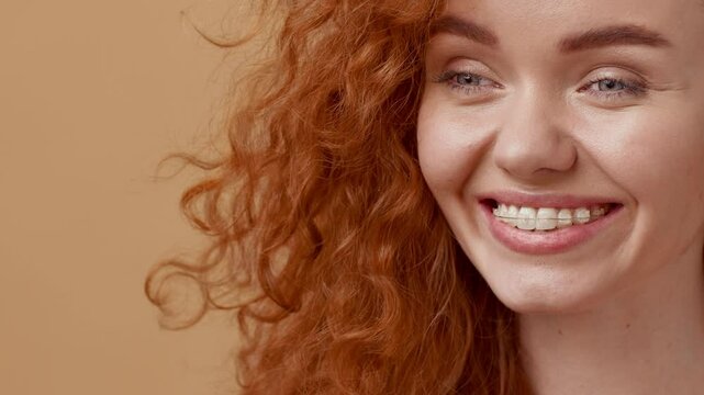 A young woman with vibrant curly red hair gazes softly, displaying a warm smile. The backdrop is a soothing orange hue, creating a relaxed and inviting atmosphere.