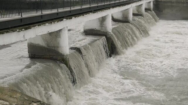Concrete dam with river flow under bridge in city.