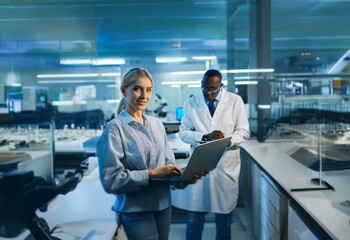 Naklejka premium Two people stand in a lab setting, a woman using a laptop and a man wearing a lab coat.