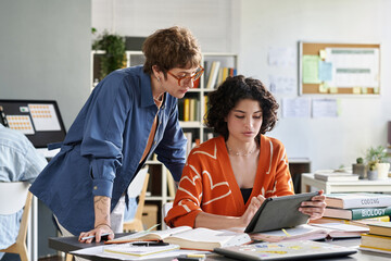Two women collaborating on a digital tablet in an office environment, surrounded by books,...