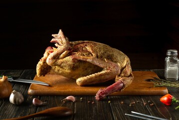 A whole duck, expertly seasoned and ready for cooking, lies on a rustic wooden cutting board surrounded by fresh ingredients. The kitchen ambiance suggests a culinary adventure