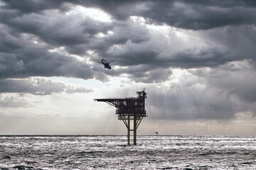 Helicopter flying towards offshore oil rig in the sea for landing, with clouds in the sky and blue...
