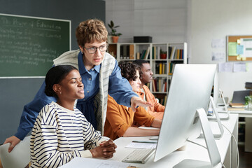 Students sitting at desks with desktop computers while teacher guiding one of them in a classroom...