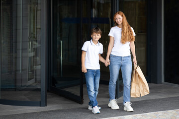 Fototapeta premium Mother and her little son standing near modern building holding hands and smiling. Mom with brown bags. Back to school shopping in supermarket with stationery. Sale. Shopping time