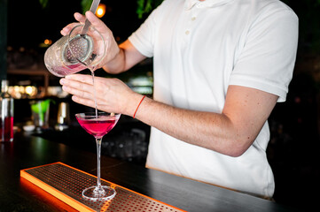 bartender in a white polo shirt pours a pink cocktail from a mixing glass into a cocktail glass at a bar. The background features various bar elements, creating a professional and inviting atmosphere.