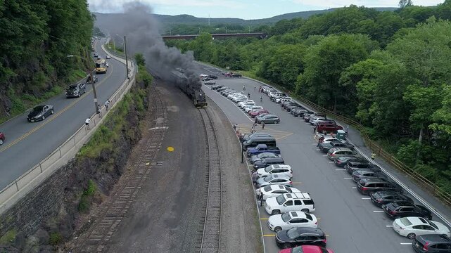 A Steam Passenger train engine emits smoke while passing along a roadway. Cars are parked nearby, and the lush landscape creates a natural backdrop on a warm summer day