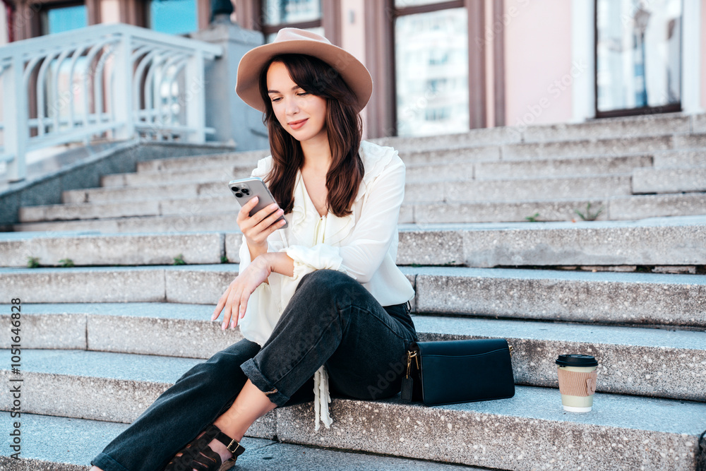 Wall mural Young beautiful smiling woman in trendy summer clothes. Carefree female posing in street in sunny day. Positive model holds smartphone, looks at cellphone screen, uses mobile apps, in hat