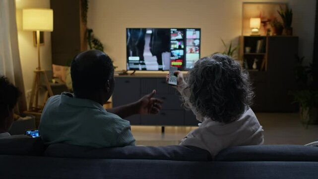 Rear view of diverse married couple sitting on sofa with kid and arguing what movie to watch on TV, taking remote control from one another, using streaming service at home