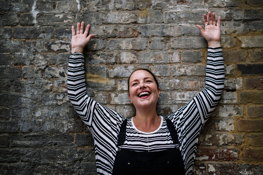 Cheerful mature woman with hands raised standing in front of wall