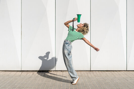 Carefree young woman shouting through megaphone in front of wall