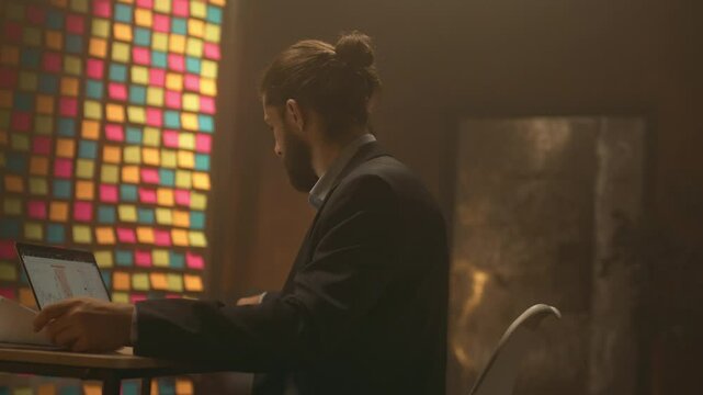 Portrait of serious businessman thinking sit at desk working with paper. Use laptop at night office. Concentrate on work. Evening light
