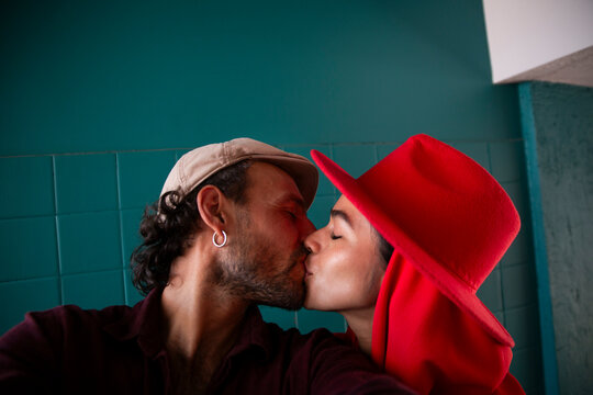 Man kissing woman wearing red hat and taking selfie in front of blue wall
