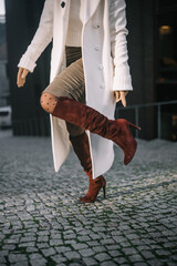 A woman in a white coat and red knee-high boots walks confidently on cobblestones
