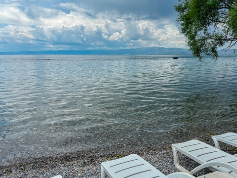 White sun loungers on pebble Beach Potpesh at the idyllic shore of Lake Ohrid, North Macedonia. Crystal clear water reflecting surrounding hills and mountains. Relaxation on summer vacation, Balkans