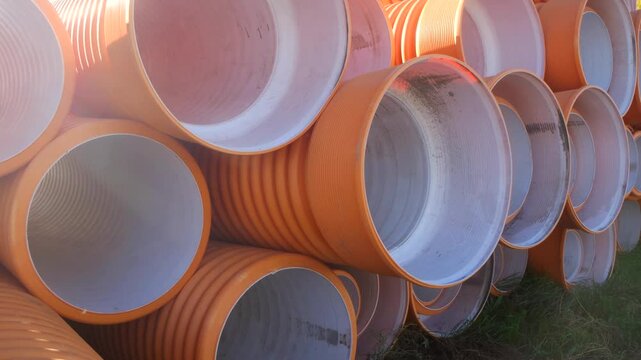 Camera panning right reveals a large stack of new, orange corrugated plastic drainage pipes at a construction site, showcasing their texture and industrial design under sunlight