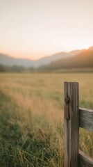 Fototapeta premium Wooden fence post in foreground with a tranquil sunset over a grassy field and mountains in the background.