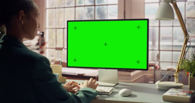 Professional Black Female Using Green Screen Monitor for Content Creation. Creative Woman working on Chroma Key Display. Modern Home Office with Warm Wooden Minimalist Interior. Over the Shoulder Shot