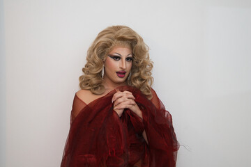 Fototapeta premium A drag queen with blonde wig and luxurious earrings and covered in red silk and on a white background with clasped hands praying imitating the virgin. Concept of lgtbqi rights and equality.