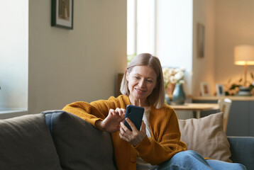 Portrait of smiling mature woman using smartphone and scrolling social media while sitting on couch...
